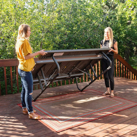 Two women setting up a Six Foot Folding Picnic Table With Classic Rugged Steel Frame Seats Eight UV Protected Top Weather and Rust Resistant Flat Fold on a deck.
