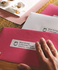 No Sweat White Mailing Address Labels For Slick Office Packing on pink envelope with hand, ideal for organizing shipments.
