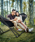 Universal Hammock Stand 600 Lb Capacity, 30 Second Foldable Setup, No Tools Or Screws Needed, Portable Camping Hammock Stand With 3 Year Warranty, Heavy Duty Steel Frame For Indoor Outdoor Use, Black in a forest setting with two people smiling.