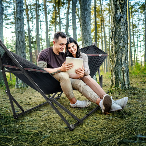 Universal Hammock Stand 600 Lb Capacity, 30 Second Foldable Setup, No Tools Or Screws Needed, Portable Camping Hammock Stand With 3 Year Warranty, Heavy Duty Steel Frame For Indoor Outdoor Use, Black in a forest setting with two people smiling.