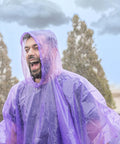 Man wearing purple rain poncho outdoors, smiling in the rain. Four Pack Disposable Rain Ponchos With Hood For Adults Clear See Through Emergency Rain Gear Lightweight And Waterproof Easy To Pack Grab And Go Perfect For Travel Outdoor Fun And Sudden Showers.