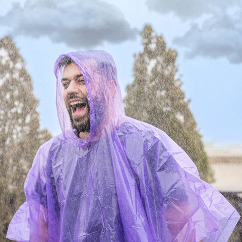 Man wearing purple rain poncho outdoors, smiling in the rain. Four Pack Disposable Rain Ponchos With Hood For Adults Clear See Through Emergency Rain Gear Lightweight And Waterproof Easy To Pack Grab And Go Perfect For Travel Outdoor Fun And Sudden Showers.