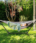 Person relaxing in a 550 lb capacity double hammock with stand and carry bag for outdoors and indoors, heavy duty two person hammock with 60x80 inch bed, desert stripe, set up in a garden.