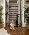 Baby sitting in front of a walk through baby and pet gate easy to use for doorways and stairways no drill pressure mount adjustable from 28 to 42 inches wide and 30 inches tall hardware mounted install on wall or banister auto close door in gray.