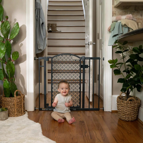 Baby sitting in front of a walk through baby and pet gate easy to use for doorways and stairways no drill pressure mount adjustable from 28 to 42 inches wide and 30 inches tall hardware mounted install on wall or banister auto close door in gray.