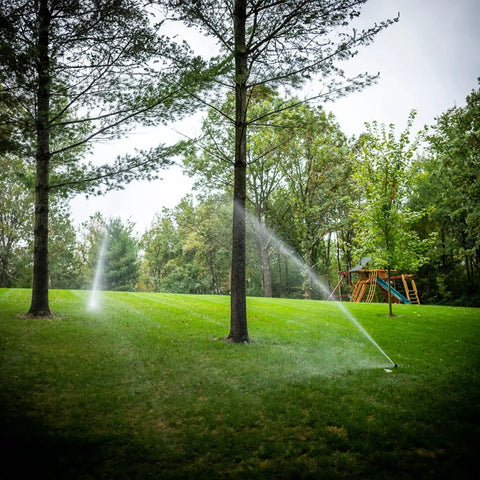 Heavy Duty Rock Solid Half Inch Zinc Impact Sprinkler 2 Pack spraying water on lush lawn between trees.