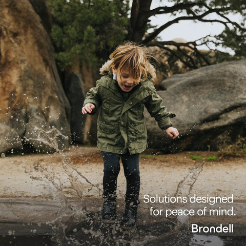 Child joyfully splashing in a puddle wearing a green jacket, with rocks and trees in the background.
