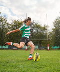 Rugby player kicking ball on field, wearing green and white stripes with goal posts in background under a cloudy sky.