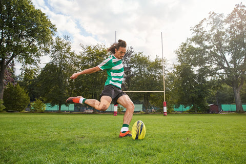 Rugby player kicking ball on field, wearing green and white stripes with goal posts in background under a cloudy sky.