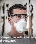 Man wearing Super Comfy N95 Particulate Respirator Pack Of 20 With 3 Panel Flat Fold Design For Easy Facial Movement Reduces Eyewear Fogging Disposable Individually Wrapped Comfortable NIOSH Approved For Smoke And Dust, with safety glasses, in a workshop.