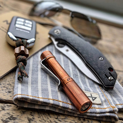 Pocket Size EDC Flashlight With Clip 92 Plus CRI Natural Light placed on a striped cloth next to a knife and keychain