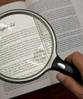 Person holding Lit 5 Inch Round Magnifier With LED 2X Magnification And 6X Bifocal Lens over an open book text.