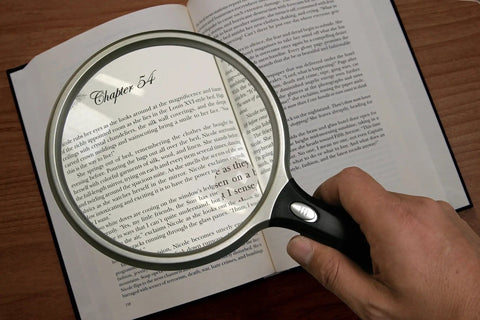 Person holding Lit 5 Inch Round Magnifier With LED 2X Magnification And 6X Bifocal Lens over an open book text.