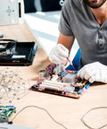 Person in gloves repairing a computer motherboard, using tools; focus on small electronic components on the workspace.