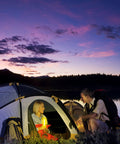 A woman inside a tent holding a Red 6 Inch Industrial Grade Light Stick while a man sits outside at dusk, with a lake and mountains in the background.