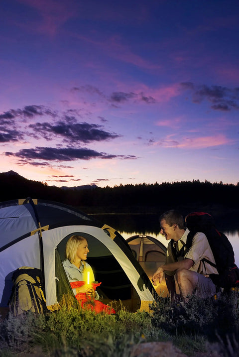 A woman inside a tent holding a Red 6 Inch Industrial Grade Light Stick while a man sits outside at dusk, with a lake and mountains in the background.