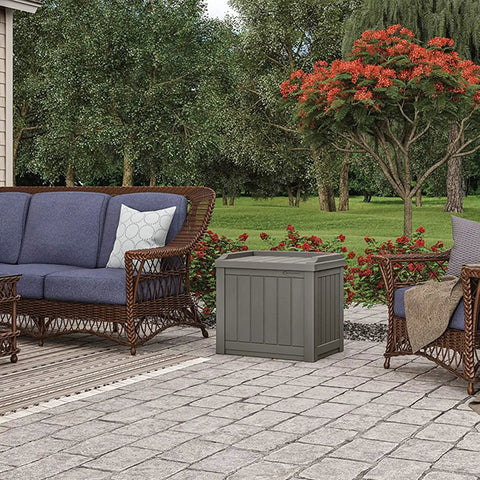 22 Gallon Outdoor Storage Box With Seat In Stone Gray Resin on a patio surrounded by wicker furniture and trees