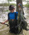 Person holding a catch of fish in a flooded forest, wearing a cap and waders, showcasing successful use of fishing tackle kit.