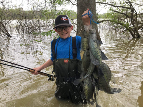 Person holding a catch of fish in a flooded forest, wearing a cap and waders, showcasing successful use of fishing tackle kit.