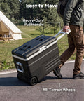 Person pulling a Smart 12V Car Fridge 59 Quarts With App Control And LCD Panel on all-terrain wheels in a camping setting.