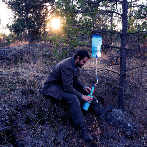 Gravity Water Filter Portable Purifier With 3L Bag And Tree Strap For Camping Hiking Emergency Prep BPA Free Survival Gear Ready Clean Water Anywhere On The Go in use, attached to a tree branch at sunset.