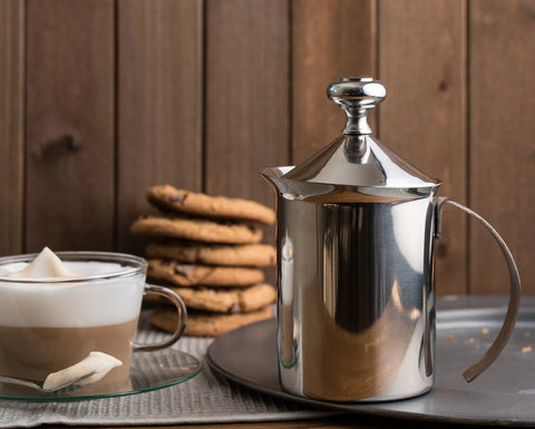 Handheld Milk Frother For Creamy Foam In Seconds, On The Go Stainless Steel Foamer next to glass cup with foam and cookies