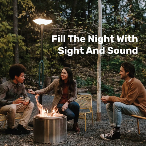 People enjoying a fire pit under Cordless Outdoor Solar Floor Lamp With Bluetooth Speaker No Wires Needed For Patios And Decks Weatherproof All Weather Outdoor Space 100 Percent Solar Powered Firefly Two Point Zero Light And Sound in a forest setting.