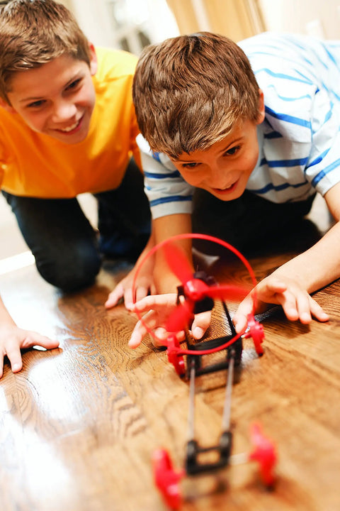 Green Science Propeller Powered Race Car Kit For Kids Ages 8 And Up Build An Epic Fast Propeller Driven Racer With Motor Wheels Frame And Battery Case To Race On Smooth Surfaces And Learn Basic Engineering With Easy Assembly. Boys race a red propeller car on wood floor.