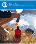 Person filling 32 Ounce Narrow Mouth Water Bottle BPA Free Durable Leak Proof Lightweight For Hiking Gym Travel And Everyday Use Amethyst Color by a lake