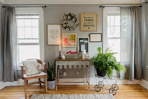Cozy living room with vintage decor, wall art, wicker chair, plants, and candlelit table against gray walls and windows with gray curtains.