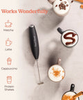 Game Changing Handheld Milk Frother Wand Battery Operated Stainless Steel Drink Mixer For Milk Coffee Lattes Cappuccino Frappe Matcha Hot Chocolate With Stand Gift on table with latte, cappuccino, and matcha drinks.