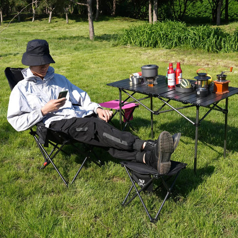 Person sitting outdoors with feet on 2 Pack 13 Inch Portable Folding Camping Stool With Carry Bag for outdoor adventures.