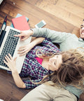 Couple relaxing on wooden floor, using a laptop together, with books and a smartphone nearby.