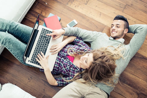 Couple relaxing on wooden floor, using a laptop together, with books and a smartphone nearby.