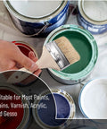 Hand holding a chip brush over green paint can with paint cans around. Six Pack Three Inch Chip Paint Brushes With Thick Natural Bristle
