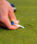 Hand placing a Premium Slick Rubberized Golf Tee With Durable Plastic Base For Low Spin Launch Assorted Colors 4 Pack into grass.