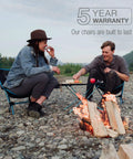 Lightweight Collapsible Camping Chair For Backpacking And Outdoors used by two people near campfire, 5-year warranty message visible