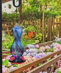 Outdoor Hand Blown Glass Hummingbird Feeder With 24 Oz Capacity And Four Feeding Ports, hanging in garden with flowers.