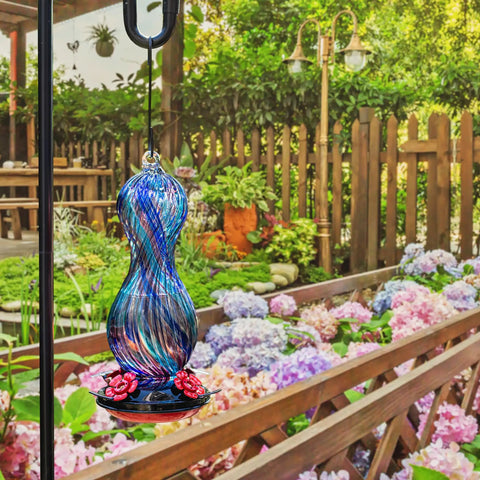Outdoor Hand Blown Glass Hummingbird Feeder With 24 Oz Capacity And Four Feeding Ports, hanging in garden with flowers.