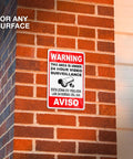 The Real Deal 24 Hour Security Camera Alert Eight By Twelve Inch Reflective Aluminum Yard Sign bold bilingual lettering mounted on brick wall.