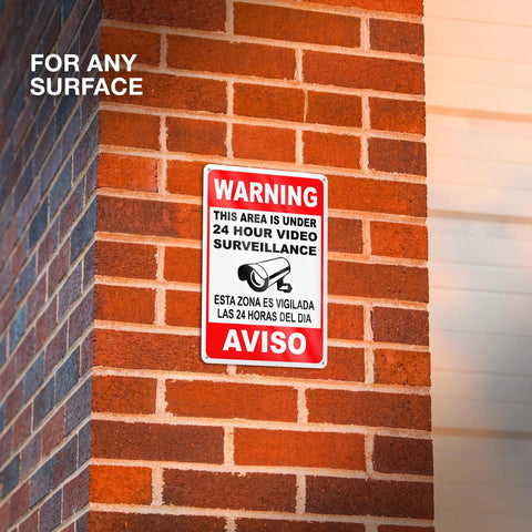 The Real Deal 24 Hour Security Camera Alert Eight By Twelve Inch Reflective Aluminum Yard Sign bold bilingual lettering mounted on brick wall.