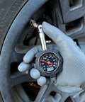 JACO Deluxe Tire Pressure Gauge, black dial, 0–100 psi, held by gloved hand near tire valve.
