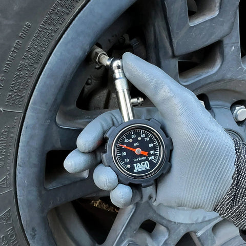 JACO Deluxe Tire Pressure Gauge, black dial, 0–100 psi, held by gloved hand near tire valve.