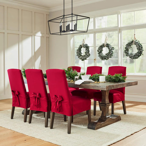 Dining room with red slipcovered chairs around wooden table, holiday wreaths on windows, and chandelier overhead.
