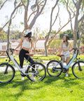 Two women chatting with Cityscape 2.0 Electric Commuter Bikes For Adults With 468Wh Removable Battery 26 Inch Wheels in a park.
