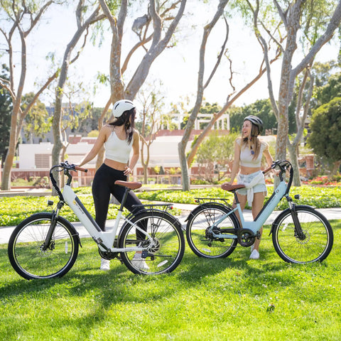 Two women chatting with Cityscape 2.0 Electric Commuter Bikes For Adults With 468Wh Removable Battery 26 Inch Wheels in a park.