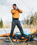 Person standing by a kayak in outdoor gear, adjusting equipment, with gear including Aquaseal FD Flexible Durable Repair Adhesive nearby