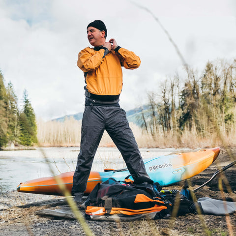 Person standing by a kayak in outdoor gear, adjusting equipment, with gear including Aquaseal FD Flexible Durable Repair Adhesive nearby