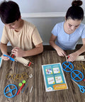 Kids assembling DIY Rubber Band Powered Car Kit For Kids 2-Pack Hands-On STEM Project with wooden parts and instructions on table