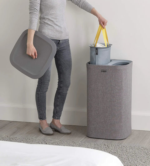 Person removing wash bag from Tota Laundry Hamper With Dual Compartments And Lid, 60 Liter Separation Basket in gray by a bed.
